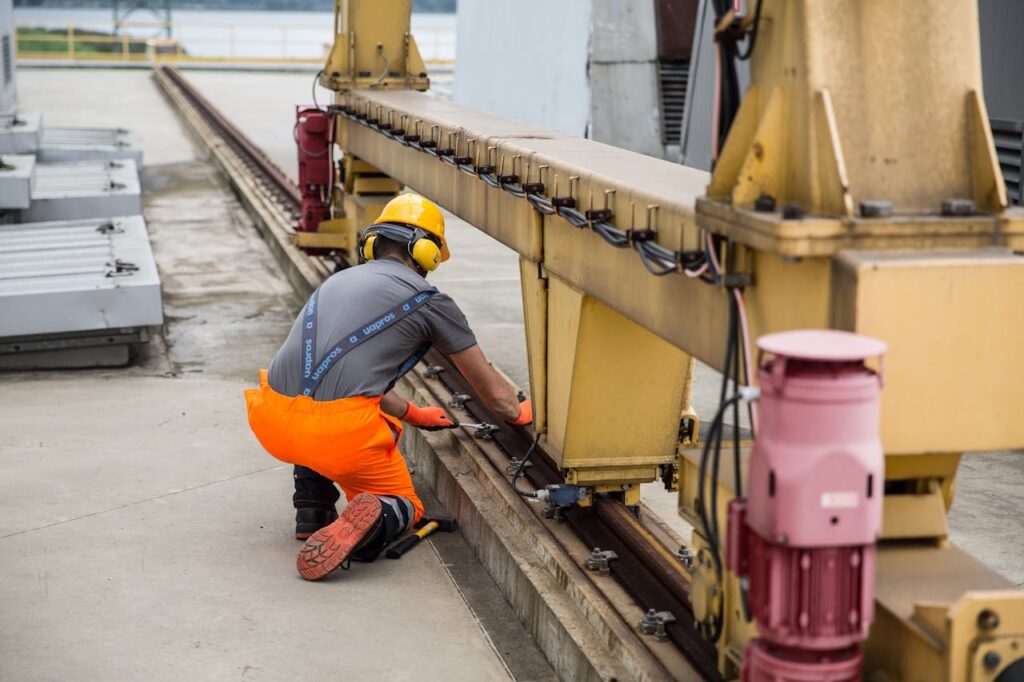A man in orange and yellow work clothes repairs a rail, focused on his task in a construction environment.